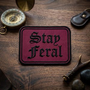 May include: A burgundy rectangular patch with the words "Stay Feral" embroidered in black gothic lettering. The patch has a black border. A compass, leather keychain, and antler are also visible on a wooden surface.