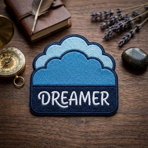 May include: A blue cloud-shaped patch with the word "DREAMER" embroidered in white. The patch has a layered design with varying shades of blue. The patch is on a wooden surface, with a compass, notebook, and lavender sprigs in the background.