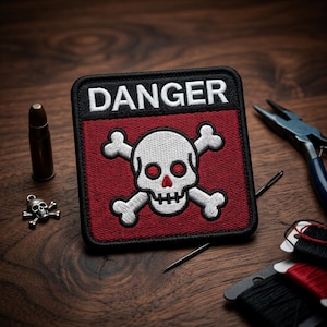 May include: A square patch with a skull and crossbones design, with the word "DANGER" in white letters on a black background. The skull is white with red eyes and nose. The patch is on a wooden surface.