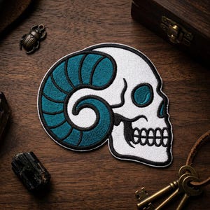 May include: Embroidered patch featuring a white skull with teal ram horns and teal eye sockets, outlined in black. The patch is on a wooden surface, with a small bronze beetle, a wooden box, and keys in the background.