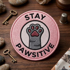 May include: A round, pink embroidered patch with the words "STAY PAWSITIVE" in black lettering. The patch features a gray cat paw with a pink paw pad in the center. The patch is on a wooden surface with keys and stones.