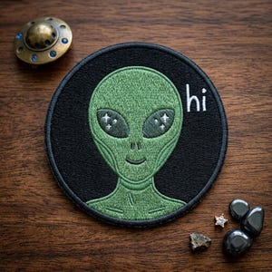 May include: Embroidered patch featuring a green alien with large eyes and the word "hi" in white. The alien is on a black circular background. A small, antique-looking metal disc and other small objects are also visible.
