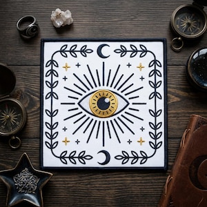 May include: Square white patch with an embroidered eye design. The eye has a gold iris and is surrounded by radiating lines, a crescent moon, and a leafy border. The patch is on a wooden surface with other mystical items.