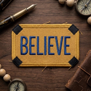 May include: A rectangular patch with the word "BELIEVE" embroidered in blue on a gold background. The patch has black corner accents and is surrounded by a gold border. The patch is on a wooden surface with a pen, compass, and journal.