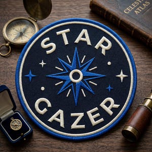 May include: A navy blue embroidered patch with the words "STAR GAZER" in white, surrounding a blue compass rose design. The patch is circular with a white border. Additional celestial elements include stars. A compass, telescope, and a small pendant are also visible.