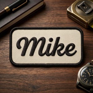 May include: A rectangular patch with the name "Mike" embroidered in black script on a cream-colored background. The patch has a black border and is set against a wooden surface, with a pen, notebook, tape measure, and watch in the background.