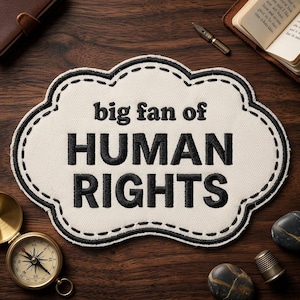 May include: A cream-colored patch with a cloud-like shape and black stitched border. The patch reads "big fan of HUMAN RIGHTS" in bold, black letters. The patch is surrounded by a compass, a small book, and other objects.