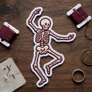 May include: Embroidered patch of a dancing skeleton in cream, burgundy, and white. The patch is on a dark wood surface with spools of burgundy thread, a small piece of fabric with needles, a penny, and a silver ring.