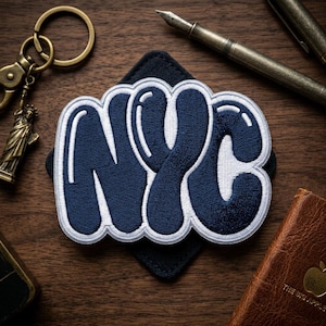 May include: Embroidered patch with the letters "NYC" in a navy blue bubble font with white trim. The patch is on a black square and surrounded by a bronze keychain with a Statue of Liberty charm, a pen, and a small leather-bound book.