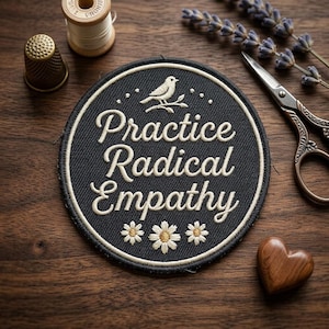 May include: A round, black embroidered patch with the words "Practice Radical Empathy" in white script. The patch features a bird on a branch and three daisy-like flowers. Other items include a thimble, thread spool, scissors, and a wooden heart.