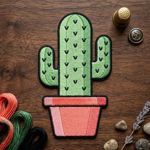 May include: Embroidered cactus patch in a terracotta pot. The cactus is light green with black details, and the pot is coral. The patch is on a wooden surface, with sewing supplies and small decorative items.