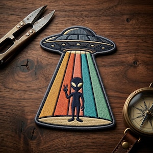 May include: Embroidered patch depicting a retro UFO with colourful beams and an alien figure giving a peace sign. The patch is in shades of blue, orange, yellow, and green. A vintage compass and scissors are also visible.