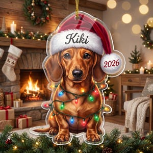 May include: A clear acrylic Christmas ornament shaped like a dachshund wearing a red Santa hat with the name Kiki printed on the brim and the year 2026 on the pom-pom. The dog is wrapped in a string of colorful, glowing Christmas lights.