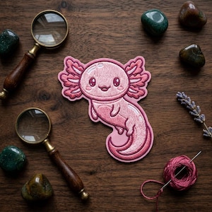 May include: A pink axolotl patch with embroidered details, featuring a smiling face and frilly gills. The patch is surrounded by magnifying glasses, gemstones, and a ball of thread on a wooden surface. A cute, whimsical design.