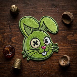 May include: A green embroidered bunny patch with a black cross for an eye and a pink heart nose. The patch is surrounded by thimbles and acorn caps on a wooden surface.