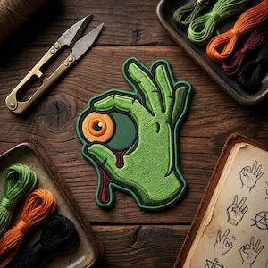 May include: Embroidered green patch featuring a hand making the "okay" gesture, holding an eyeball with orange and brown details. The eyeball has a red drip. Also shown are embroidery floss, scissors, and a notebook with hand-drawn illustrations.