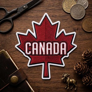 May include: A red maple leaf patch with the word "CANADA" in white lettering. The patch has a black and white border. The image also includes Canadian coins, scissors, a small brass moose figurine, and pine cones, all arranged on a wooden surface.