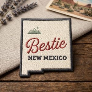May include: Embroidered patch in the shape of New Mexico with the word "Bestie" in red script and "NEW MEXICO" in black block letters. A small mountain graphic is above the word "Bestie". The patch has a black border and is on a beige background.