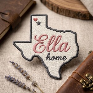 May include: Embroidered patch in the shape of Texas, with the name "Ella" in pink script and the word "home" below. A small heart and star are embroidered in the upper corner. The patch has a gray border and is on a beige background.