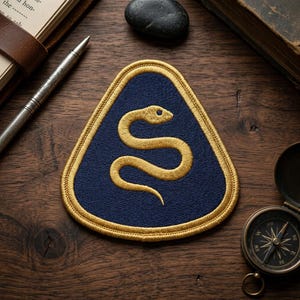 May include: A navy blue, triangular patch with a gold embroidered snake design. The patch has a gold border and is set against a wooden background with a pen, book, and compass.