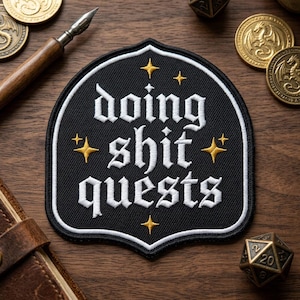 May include: Black patch with white embroidered text "doing shit quests" and gold star accents. The patch has a white border and a unique shape. Other items include coins, a pen, and a die.