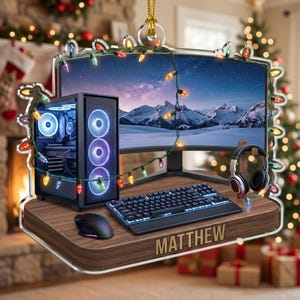 May include: A flat acrylic ornament featuring a gaming setup with a computer tower, curved monitor displaying a snowy mountain landscape, keyboard, mouse, and headphones on a wooden desk, wrapped in colorful Christmas lights with the name MATTHEW.