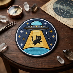 May include: Embroidered patch featuring a black cat being beamed up by a UFO. The patch has a navy blue background with yellow stars and a blue and yellow beam of light. The UFO is gray and blue.