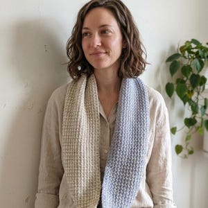 Peut inclure: Écharpe infinie bicolore avec un motif en tricot gaufré. Un côté est beige clair, l'autre bleu doux. L'écharpe est enroulée autour du cou, mettant en valeur la texture et le contraste des couleurs. Le fond est un mur neutre.