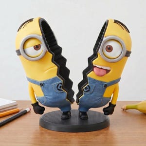 Puede incluir: Figura dividida de Minion amarillo con peto azul y guantes negros, sobre una base negra. Un lado tiene una expresión triste, el otro está feliz. Un plátano y lápices están sobre la superficie de madera.