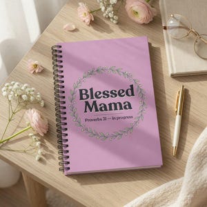 Puede incluir: Un cuaderno espiral lila con el texto "Blessed Mama" dentro de una corona floral y "Proverbs 31 - in progress" debajo. Un bolígrafo dorado, gafas y flores están sobre una mesa de madera. Un objeto útil.