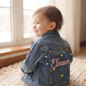Puede incluir: Una chaqueta de mezclilla azul de tono medio con el nombre Eleanor bordado en letra cursiva rosa en la parte posterior. La chaqueta está decorada con pequeños corazones bordados de colores en tonos amarillo, rosa, turquesa y naranja.