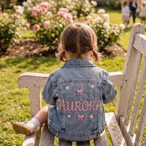 Puede incluir: Una chaqueta de mezclilla azul claro con el nombre Aurora bordado en letra cursiva rosa en la parte posterior. El diseño incluye pequeños lazos rosas, flores blancas y corazones rosas bordados alrededor del nombre.