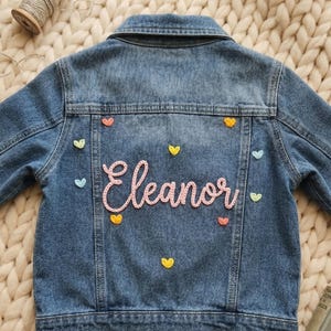 Puede incluir: Chaqueta vaquera azul con el nombre "Eleanor" en escritura rosa, rodeada de pequeños apliques en forma de corazón de colores. Chaqueta clásica para niños.