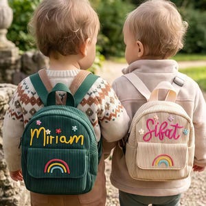 Puede incluir: Dos niños con mochilas pequeñas. Una es de pana verde con el nombre "Miriam" en amarillo, un arcoíris y detalles de flores. La otra es beige con el nombre "Sifat" en rosa, un arcoíris y detalles de flores.