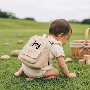 Puede incluir: Una mini mochila de pana beige con la palabra Joy bordada en letra cursiva negra, decorada con pequeñas flores blancas y un icono de sol amarillo en el bolsillo frontal. La mochila incluye correas ajustables y una pequeña etiqueta de marca.