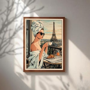 May include: A framed art print depicts a woman in a towel and sunglasses, sipping coffee on a balcony overlooking the Eiffel Tower. A croissant and newspaper rest on the table. The artwork has a vintage comic book style.