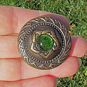 May include: A round, antique-style button with a brass finish and a central green glass stone. The button features intricate embossed patterns and a decorative border, perfect for sewing or crafting projects.