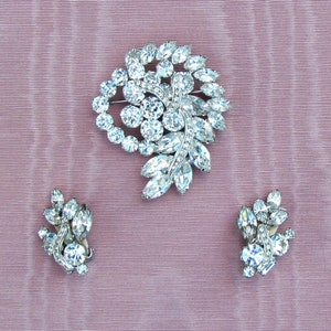 May include: A set of vintage rhinestone jewelry featuring a large brooch and a pair of earrings. The brooch is shaped like a cluster of leaves and flowers, while the earrings are smaller and more delicate.