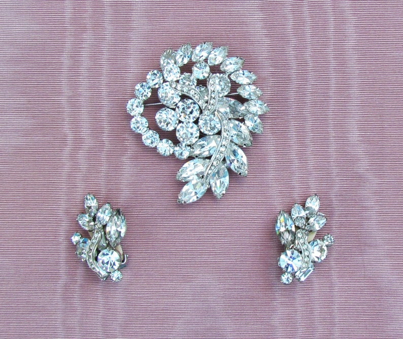 May include: A set of vintage rhinestone jewelry featuring a large brooch and a pair of earrings. The brooch is shaped like a cluster of leaves and flowers, while the earrings are smaller and more delicate.