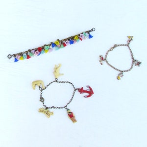 May include: Three charm bracelets. The first bracelet has a chain with colorful bell charms. The second bracelet has a chain with a red anchor, a white elephant, a white bear, a white bridge, and a red and white figure charm. The third bracelet has a chain with colorful flower charms.