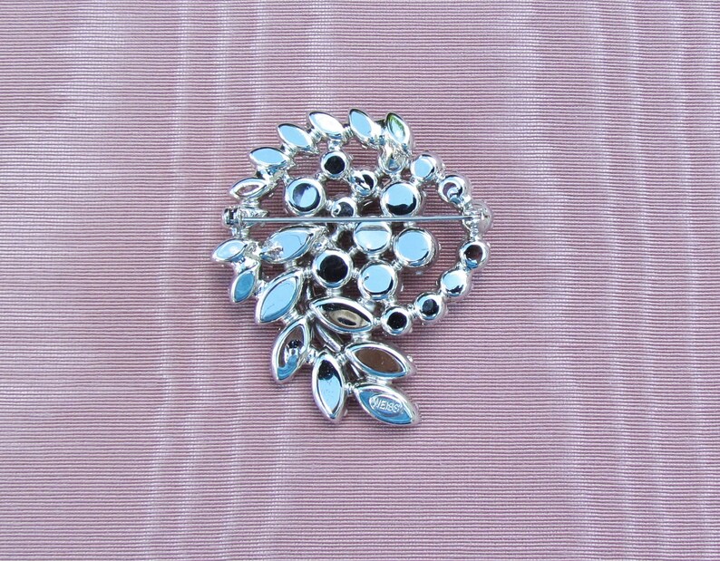May include: A silver-toned brooch with a swirl design featuring clear and dark gray rhinestones. The brooch is marked "WIESS" on the back.