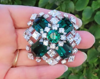 Op de afbeelding: Een broche met een grote centrale ronde smaragdgroene steen, omringd door vier rechthoekige smaragdgroene stenen. Het sieraad is versierd met talloze kleinere heldere gefacetteerde stenen in een geometrisch patroon, gezet in zilverkleurig metaal.