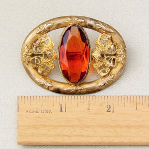 Op de afbeelding: Vintage ovale broche met een grote, gefacetteerde, oranjerode edelsteen in het midden. Het goudkleurige metalen frame heeft sierlijke filigraan- en bloemendetails. De broche is ongeveer 6,35 cm lang.