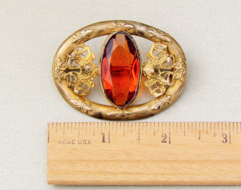 Op de afbeelding: Vintage ovale broche met een grote, gefacetteerde, oranjerode edelsteen in het midden. Het goudkleurige metalen frame heeft sierlijke filigraan- en bloemendetails. De broche is ongeveer 6,35 cm lang.
