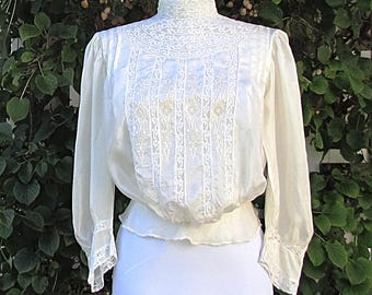 May include: Cream-colored vintage blouse with long sleeves and a high, detailed collar. The blouse features intricate lace and embroidered detailing on the bodice and cuffs. The fabric appears to be lightweight, with a gathered waistline. The blouse is displayed on a mannequin.