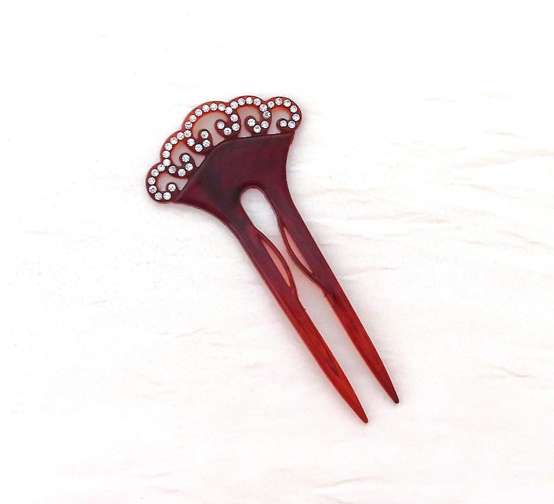 Puede incluir: Una horquilla para el cabello de color rojo oscuro con una parte superior decorativa adornada con peque&ntilde;os y brillantes pedrer&iacute;a. El accesorio para el cabello presenta un dise&ntilde;o de dos puntas, ideal para asegurar el cabello en un mo&ntilde;o. La parte superior tiene un borde festoneado.