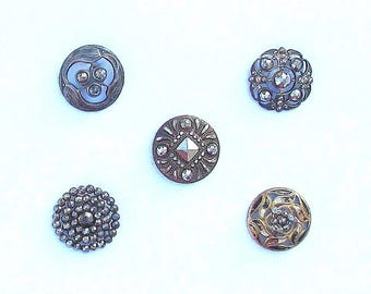 May include: Five antique buttons in various designs. The buttons are silver and gold-toned, with intricate details. One button features a floral design, while others have geometric or beaded patterns. The buttons are arranged on a white background.