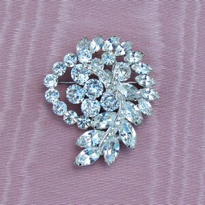May include: A silver brooch with a swirl design featuring clear rhinestones. The brooch is set against a pink fabric background.