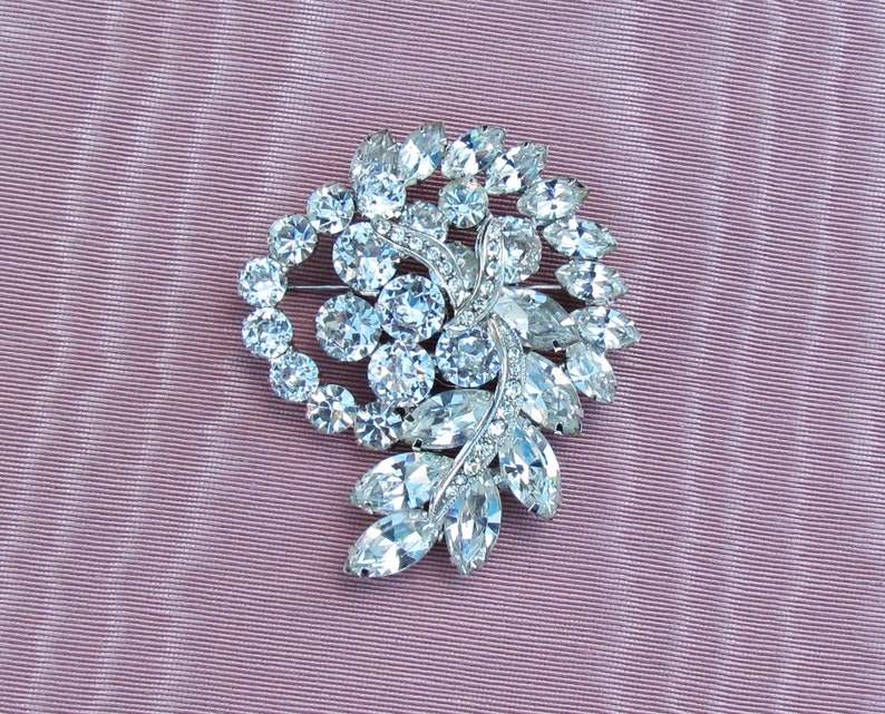 May include: A silver brooch with a swirl design featuring clear rhinestones. The brooch is set against a pink fabric background.