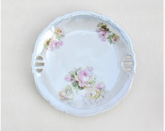 May include: A white porcelain serving dish with two handles and a floral design featuring pink and yellow roses. The dish has a scalloped edge and a delicate floral pattern.
